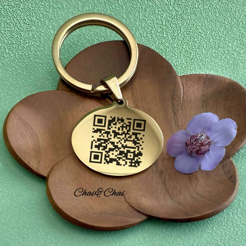 May include: A gold-coloured keyring with a round tag featuring a QR code. The keyring is attached to a wooden, flower-shaped coaster. The text "Chai & Chai" is printed on the coaster. A purple flower with a dark centre is also present.
