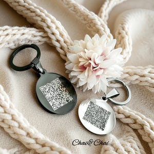 May include: Two keychains with QR codes. One is black and oval-shaped, the other is silver and round. Both have a metal ring for keys. A white and pink flower and a cream-colored rope are in the background. The text "Chai & Chai" is at the bottom.
