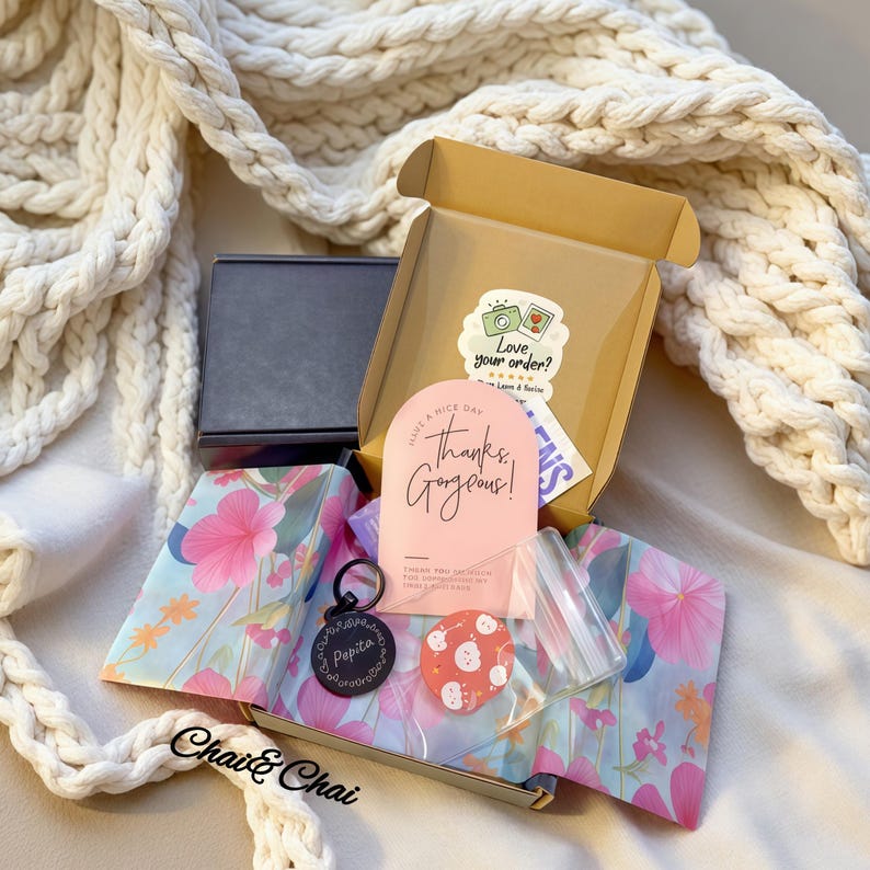 May include: A cardboard box with a sticker and a pink card reading "Thanks, Gorgeous!". Inside are a keychain, a red and white item, and items wrapped in floral paper. The brand name "Chai & Chai" is visible. The box is open on a white surface.