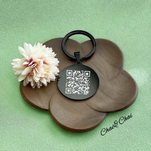 May include: A black round keychain with a QR code, attached to a black ring. The keychain rests on a brown wooden tray, next to a cream and burgundy flower. The background is a light green colour. The text "Chai & Chai" is visible on the tray.