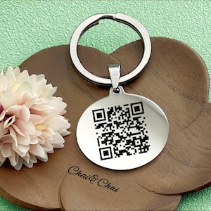 May include: A silver-colored keychain with a round pendant featuring a QR code. The keychain is attached to a wooden, cloud-shaped tray. The text "Chai & Chai" is printed on the tray. A light pink flower is on the left side.