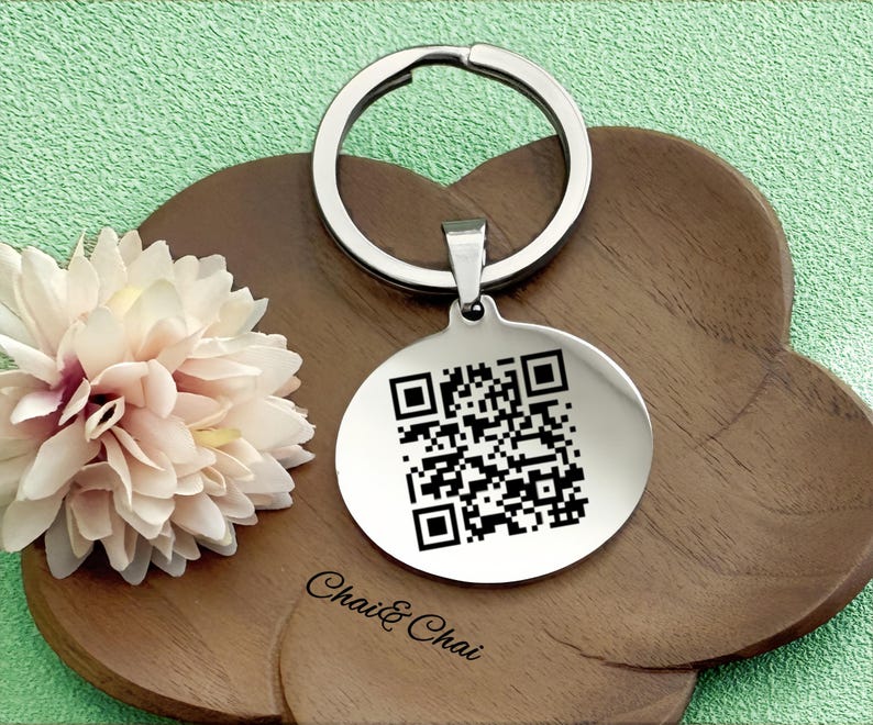 May include: A silver keychain with a circular pendant displaying a QR code. The keychain is on a wooden, cloud-shaped tray. The text "Chai & Chai" is printed on the tray. A light pink flower is on the left side.