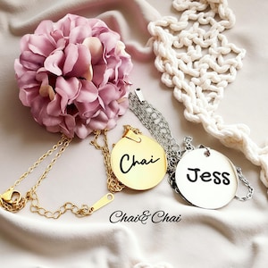 May include: Two personalized pendant necklaces. One is gold-toned with the name "Chai" in black script, the other is silver-toned with "Jess". Both are on delicate chains. The image includes a pink flower and the text "Chai & Chai".