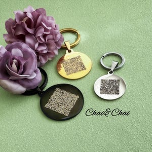 May include: Three circular keyrings in gold, silver, and black, each featuring a QR code design. The keyrings are presented with purple fabric flowers on a green backdrop. The text "Chai & Chai" is also visible.