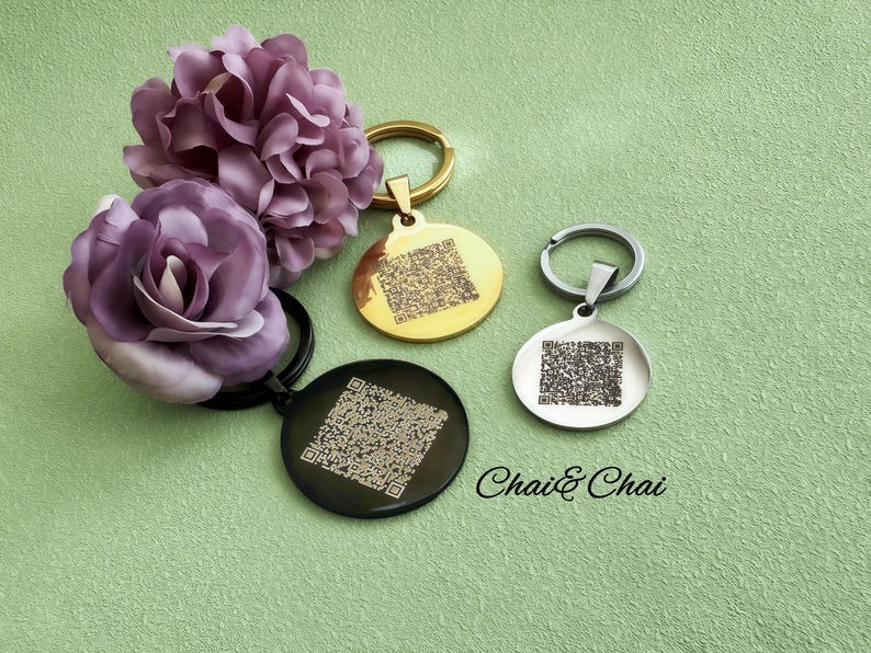 May include: Three circular keyrings in gold, silver, and black, each featuring a QR code design. The keyrings are presented with purple fabric flowers on a green backdrop. The text "Chai & Chai" is also visible.
