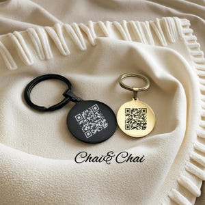 May include: Two round keychains, one black and one gold, each featuring a QR code design. The keychains are displayed on a cream-colored, textured fabric. The text "Chai & Chai" is printed below the keychains.