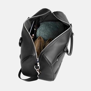 Pu&ograve; includere: Borsa da viaggio nera con cerniera argentata, aperta per rivelare il contenuto. All'interno ci sono scarpe marroni, un tessuto verde fantasia e accessori. La borsa ha una tracolla e manici.