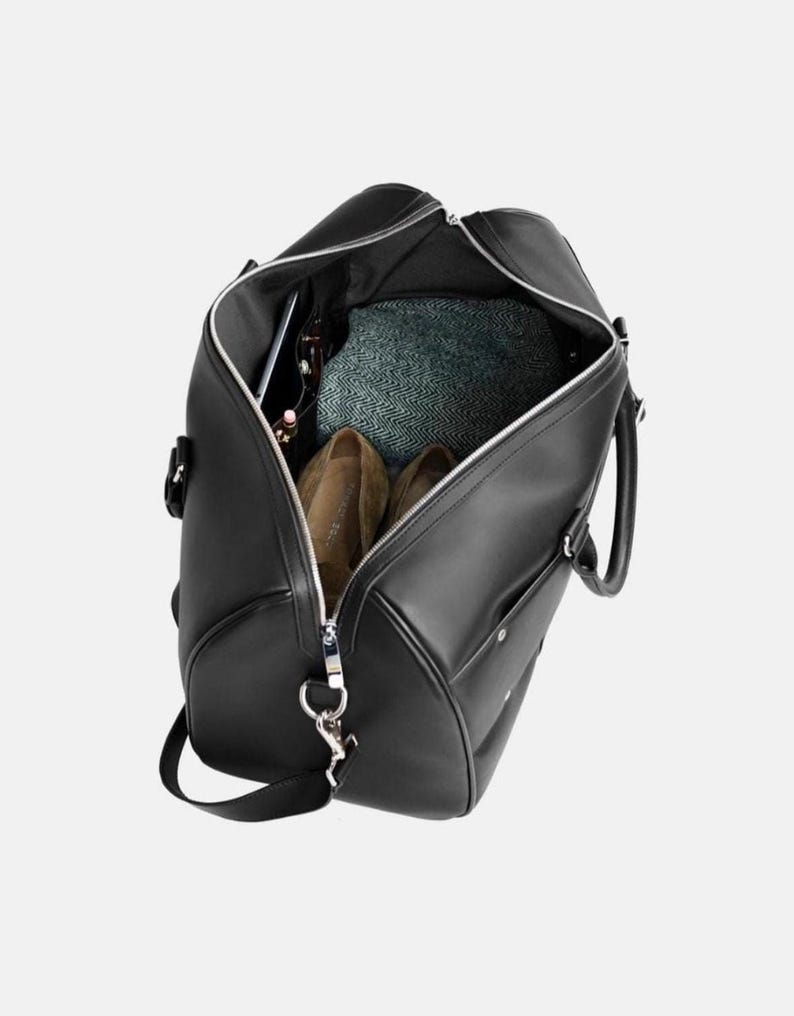 Pu&ograve; includere: Borsa da viaggio nera con cerniera argentata, aperta per rivelare il contenuto. All'interno ci sono scarpe marroni, un tessuto verde fantasia e accessori. La borsa ha una tracolla e manici.