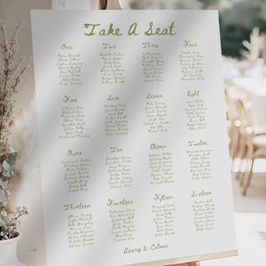 May include: A white wedding seating chart on an easel with the words "Take a Seat" in green script. The chart lists names in numbered columns, with the names Avery & Calvin at the bottom.