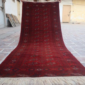 Puede incluir: Una alfombra alargada de forma rectangular con un fondo rojo intenso y patrones geométricos oscuros y motivos florales repetidos. La alfombra presenta un diseño denso y complejo en toda su superficie y termina con flecos de color crema.