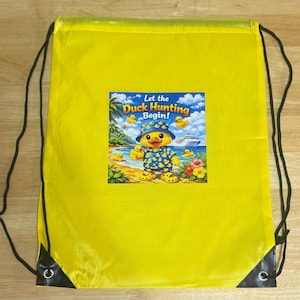 May include: Yellow drawstring backpack with a colorful graphic of a duck wearing a hat and shirt, with the text "Let the Duck Hunting Begin!". The graphic includes ducks, a beach scene, and a cruise ship.