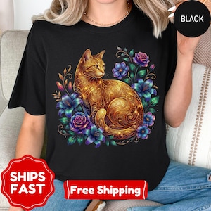Może przedstawiać: Czarny t-shirt z ilustracją złotego kota otoczonego fioletowymi różami i niebieskimi kwiatami. Kot ma skomplikowane złote wzory. Słowo "BLACK" znajduje się w czarnym okręgu w prawym górnym rogu.