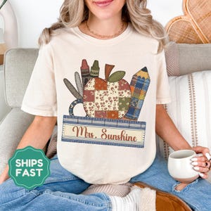 Könnte beinhalten: Cremefarbenes T-Shirt mit einem Patchwork-Apfel-Design, Buntstiften, einer Schere und einem Bleistift. Unter dem Apfel befindet sich ein blaues Lineal mit dem Text "Mrs. Sunshine". Das Shirt ist ein bequemer, lässiger Stil.