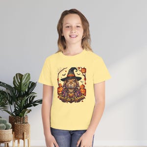 May include: A pale yellow short-sleeve t-shirt featuring a graphic print of a brown rabbit wearing a black witch hat and a purple patterned cape. The design includes orange jack-o'-lanterns, bats, and the text "Too Cute To Spook" in gold.