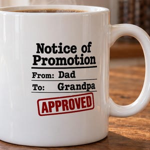 May include: A white ceramic mug featuring black text that reads Notice of Promotion, From: Dad, To: Grandpa, with a red rectangular stamp graphic below that says APPROVED. The mug has a standard handle and is filled with a dark liquid beverage.