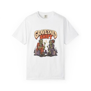 May include: A white short-sleeve t-shirt featuring a graphic print of two zombies sitting on tombstones in a graveyard, drinking from small cups. Above them, the text GRAVEYARD SHIFT is printed in a bold, orange, and cream-coloured font.