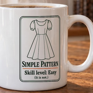 May include: A white ceramic mug featuring a dark green graphic of a dress. Below the dress, the text reads "SIMPLE PATTERN" in green, followed by "Skill level: Easy" and "(It is not.)" in smaller green text inside a rectangular border.
