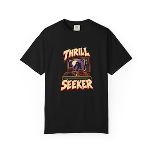 Peut inclure: Un t-shirt noir à manches courtes avec un motif représentant deux personnes assises sur un canapé devant une télévision affichant la silhouette d'une créature sombre. Le texte THRILL SEEKER est imprimé en police dentelée claire au-dessus et en dessous.