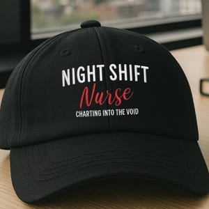 May include: A black baseball cap featuring the white text NIGHT SHIFT and CHARTING INTO THE VOID with the word Nurse in red script. Above the cap, a graphic displays the text AVAILABLE IN 6 COLORS with circular swatches for black, navy, spruce, dark grey, khaki, and stone.