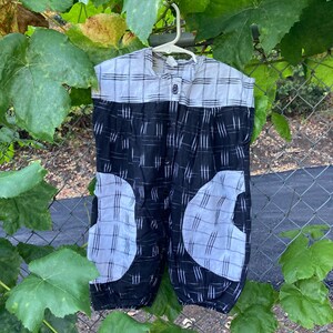 May include: A black and white patterned romper hangs on a white hanger against a backdrop of green leaves and a chain-link fence. The romper features a button-up front and pockets with a contrasting white pattern. The fabric has a textured appearance.