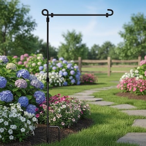 May include: A black metal garden flag stand with a curved top and a hook for displaying flags. The stand is in a garden setting with colorful flowers and green grass. The stand is approximately 36 inches tall.