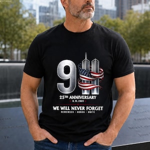 May include: A black short-sleeved t-shirt featuring a graphic of the number 9 and the Twin Towers wrapped in an American flag. Text reads 25TH ANNIVERSARY 9.11.2001 WE WILL NEVER FORGET REMEMBER HONOR UNITE in white and red lettering on the front.
