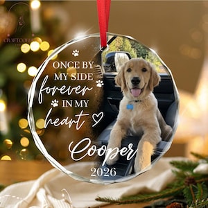 May include: A round, clear acrylic ornament featuring a photo of a golden retriever puppy sitting in a car. The text reads: ONCE BY MY SIDE forever IN MY heart Cooper 2026. The ornament is suspended by a red ribbon and includes paw print icons.