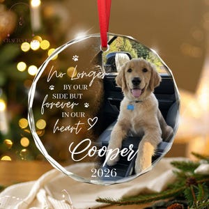May include: A circular glass ornament featuring a photo of a golden retriever puppy sitting in a car. The text reads: No Longer BY OUR SIDE BUT forever IN OUR heart Cooper 2026. It includes a red ribbon for hanging and small paw print icons.