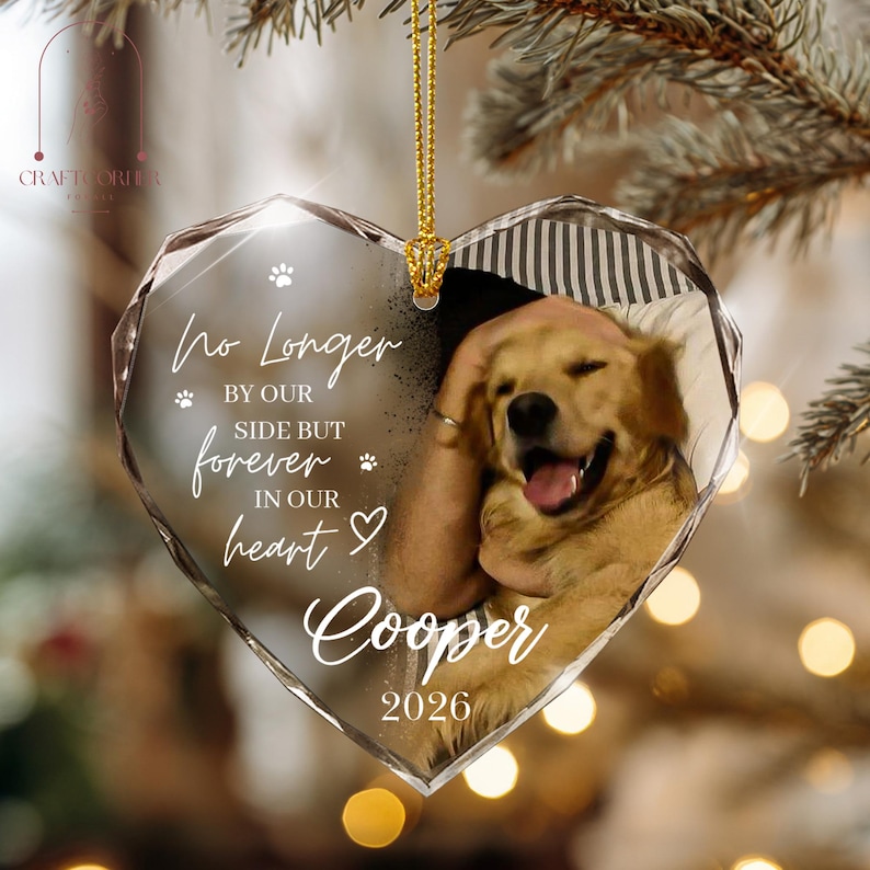 May include: A heart-shaped clear acrylic ornament featuring a golden retriever photograph, white text reading No Longer BY OUR SIDE BUT forever IN OUR heart Cooper 2026, small paw prints, and a gold hanging cord, suspended against a blurred background.