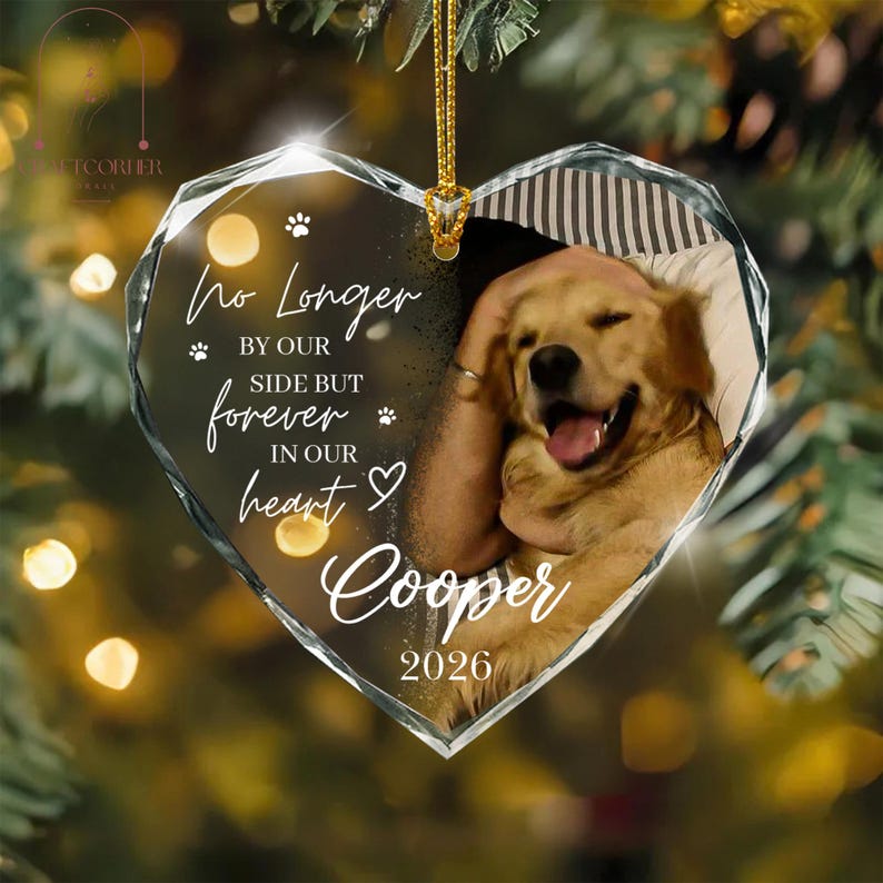 May include: A heart-shaped glass ornament featuring a golden retriever photograph, the text "No Longer BY OUR SIDE BUT forever IN OUR heart Cooper 2026", and small paw print icons. The ornament hangs from a gold cord against a blurred festive background.
