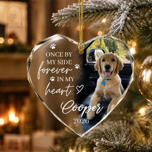 May include: A heart-shaped clear acrylic ornament featuring a photo of a golden retriever sitting in a car. The text reads: ONCE BY MY SIDE forever IN MY heart Cooper 2026, accompanied by paw print and heart icons. It hangs from a gold cord.