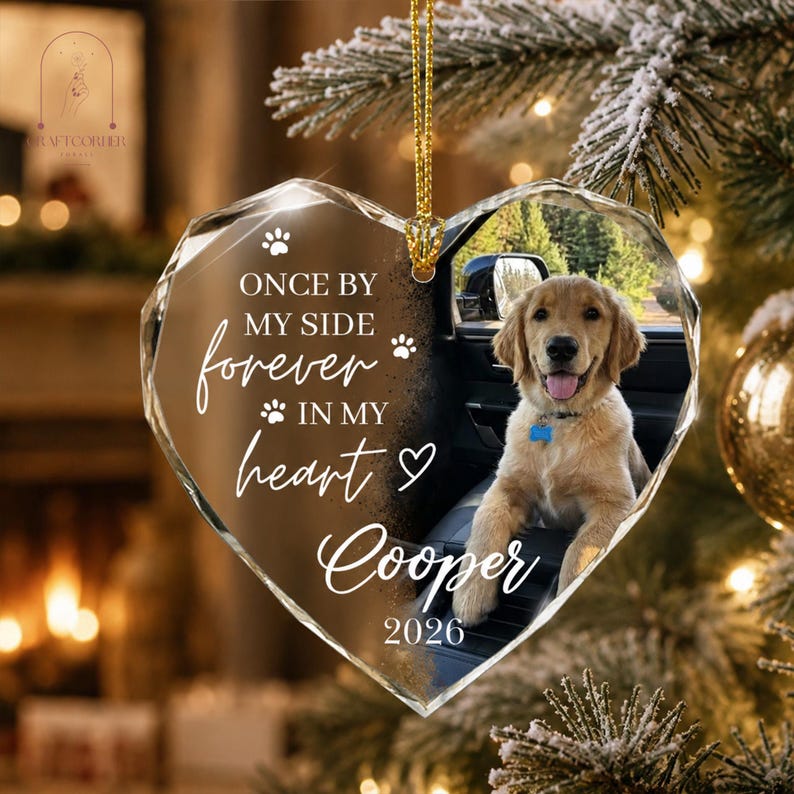 May include: A heart-shaped clear acrylic ornament featuring a photo of a golden retriever sitting in a car. The text reads: ONCE BY MY SIDE forever IN MY heart Cooper 2026, accompanied by paw print and heart icons. It hangs from a gold cord.