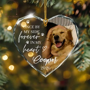 May include: A heart-shaped clear acrylic ornament featuring a golden retriever photograph, white paw prints, and the text: ONCE BY MY SIDE forever IN MY heart Cooper 2026. The ornament hangs from a gold cord against a blurred, festive light background.