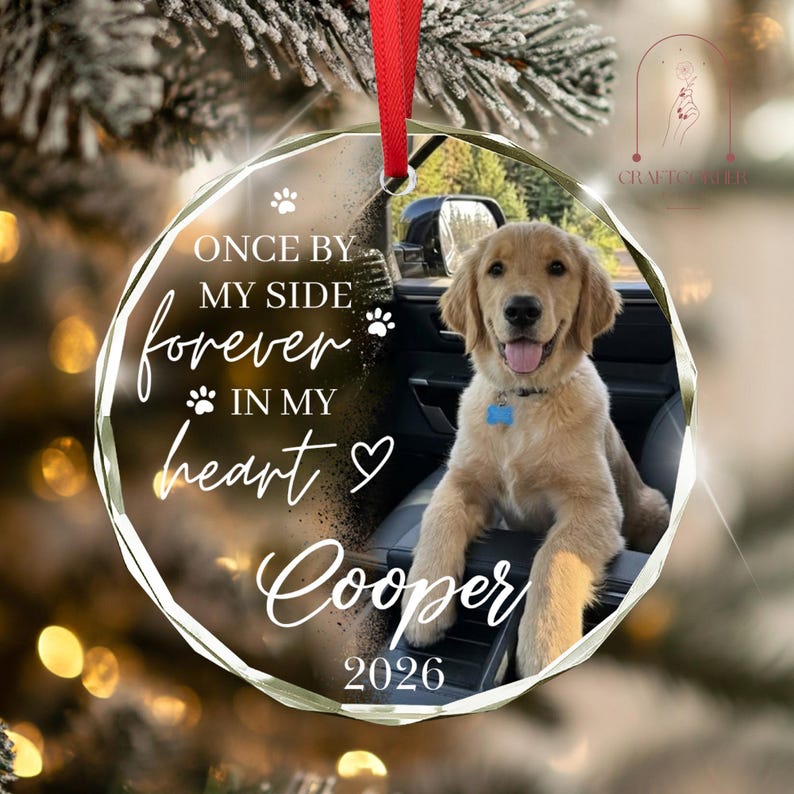 May include: A round, clear acrylic ornament with a red ribbon, featuring a photo of a golden retriever sitting in a car. The text reads: ONCE BY MY SIDE forever IN MY heart Cooper 2026, accompanied by paw print and heart icons.
