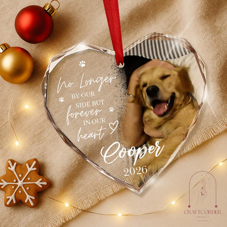 May include: A heart-shaped glass ornament with a red ribbon, featuring a photo of a golden dog and the text: No Longer BY OUR SIDE BUT forever IN OUR heart Cooper 2026. The design includes small paw prints and a heart icon on a clear surface.
