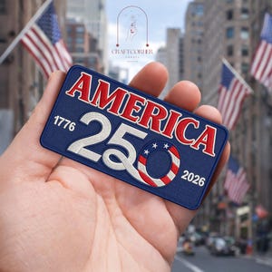 May include: A navy blue embroidered patch with the word "AMERICA" in red, the number "250" in white, and the years "1776" and "2026". The patch features a design with red and white stripes and stars.