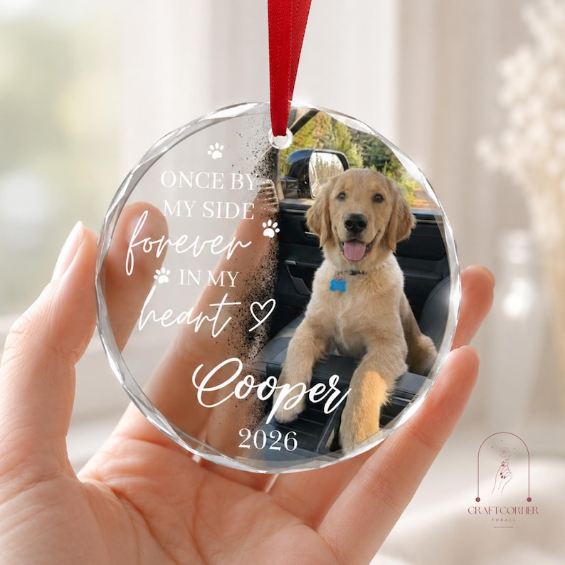 May include: A round, clear acrylic ornament featuring a photo of a golden retriever puppy sitting in a car. The text reads: ONCE BY MY SIDE forever IN MY heart Cooper 2026. It includes paw print and heart icons and hangs from a red ribbon.