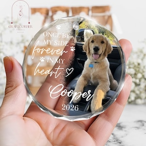 May include: A round, faceted glass ornament featuring a photograph of a golden retriever puppy sitting in a car. The text reads: ONCE BY MY SIDE forever IN MY HEART, Cooper, and 2026, accompanied by small paw print and heart icons.