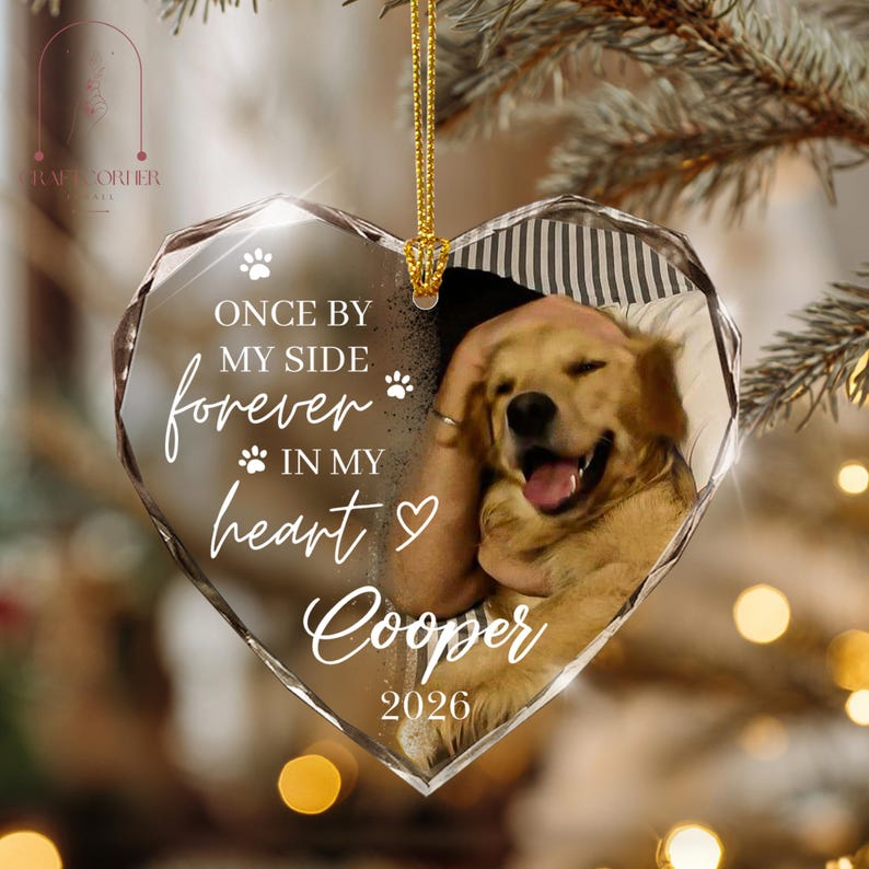 May include: A heart-shaped glass ornament featuring a photograph of a golden dog. The text reads: ONCE BY MY SIDE forever IN MY HEART Cooper 2026, accompanied by paw print and heart icons. The ornament hangs from a gold cord against a blurred background.