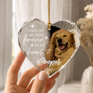 May include: A heart-shaped clear acrylic ornament with faceted edges, featuring a photo of a golden dog and the text "ONCE BY MY SIDE forever IN MY heart Cooper 2026" with paw print icons. It hangs from a gold cord for display purposes.