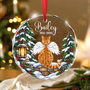 May include: A round, faceted glass ornament featuring a ginger cat with white angel wings sitting on a stone wall. The design includes snow-covered pine trees, red berries, a lantern, and the text "Bailey 2015-2026" in white script.