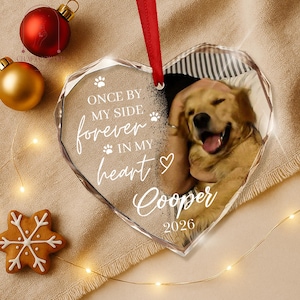May include: A heart-shaped clear acrylic ornament featuring a photo of a golden retriever, a red ribbon hanger, and white text that reads: ONCE BY MY SIDE forever IN MY heart Cooper 2026, accented with small paw prints and a heart symbol.