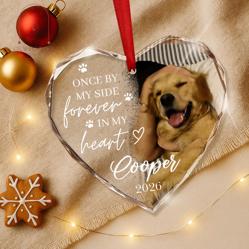 May include: A heart-shaped clear acrylic ornament featuring a photo of a golden retriever, a red ribbon hanger, and white text that reads: ONCE BY MY SIDE forever IN MY heart Cooper 2026, accented with small paw prints and a heart symbol.