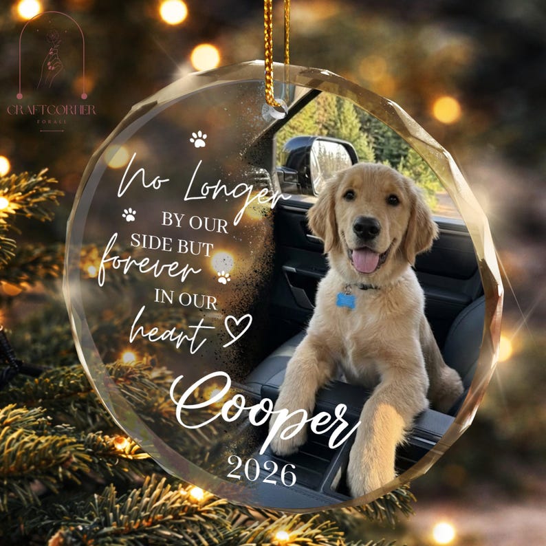 May include: A circular glass ornament featuring a golden retriever puppy sitting in a car. The text reads: No Longer BY OUR SIDE BUT forever IN OUR HEART Cooper 2026. The design includes small paw prints and a heart icon, suspended by a gold cord.