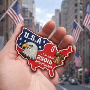 May include: A red, white, and blue embroidered patch in the shape of the USA. It features a bald eagle, the text "U.S.A.", and "250th Anniversary 1776-2026". The patch is held in a hand.
