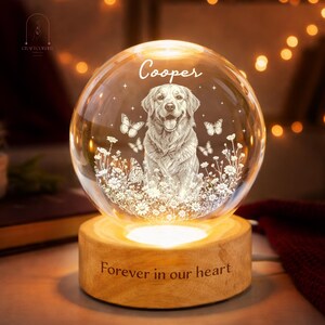 May include: A spherical glass night light featuring an etched golden retriever surrounded by daisies and butterflies. The name Cooper is engraved above the dog. The glass sphere rests on a cylindrical wooden base with the text Forever in our heart.