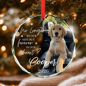 May include: A circular glass ornament featuring a photo of a golden retriever puppy sitting in a car. The text reads: No Longer BY OUR SIDE BUT forever IN OUR heart Cooper 2026. It includes a red ribbon for hanging and small paw print icons.