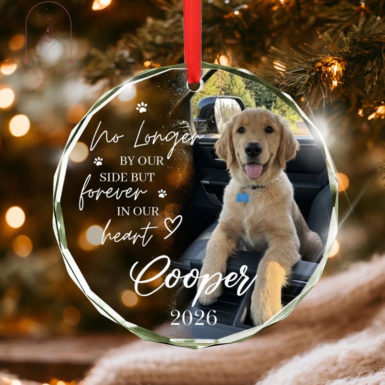 May include: A circular glass ornament featuring a photo of a golden retriever puppy sitting in a car. The text reads: No Longer BY OUR SIDE BUT forever IN OUR heart Cooper 2026. It includes a red ribbon for hanging and small paw print icons.