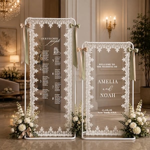 May include: Two rectangular white metal frames holding clear acrylic panels decorated with white lace trim and sage green ribbons. The left panel lists names under "OUR FAVORITE people," and the right panel displays "WELCOME TO THE WEDDING OF AMELIA and NOAH 13.06.26 NEW YORK, USA."
