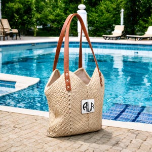 Op de afbeelding: Een beige geweven draagtas met bruine leren handvatten en een wit monogram patch. De tas heeft een uniek open ontwerp en staat bij een zwembad. De handvatten zijn ongeveer 61 cm lang.